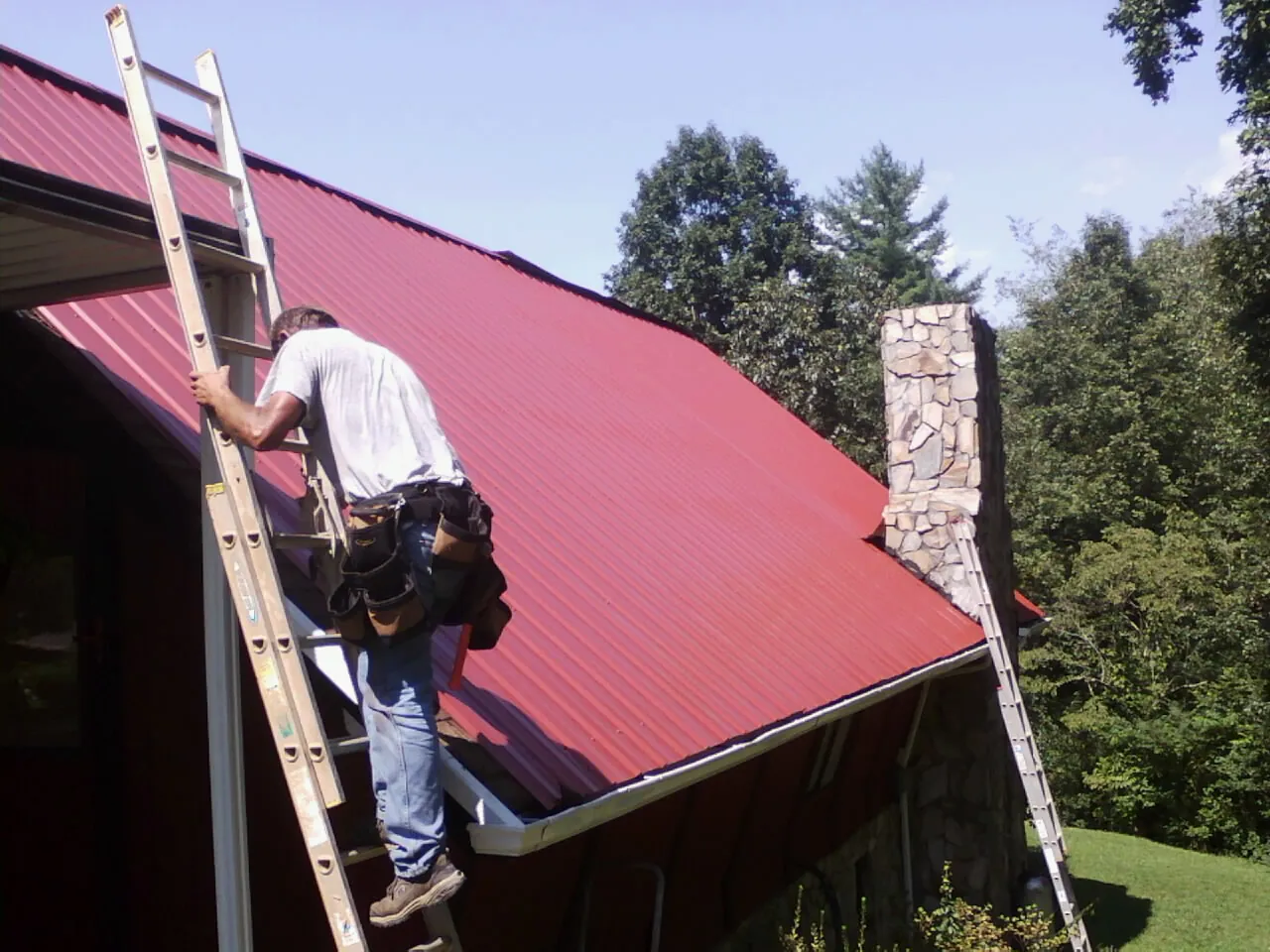 Roofer on a ladder beside a red metal roof and stone chimney — Soffit & Fascia Repair by Roofing Brothers in Theresa