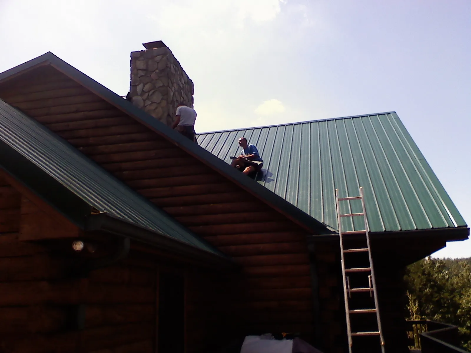 Roofers installing green metal panels beside a stone chimney on a log home in Theresa